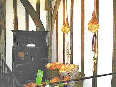 The buttery of the old hall, hams and onions hanging from the beams beside a black range and baskets of bread.