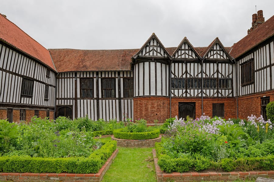 The south courtyard of Gainsborough Old Hall on an overcast day. Two timber-framed ranges meet at a right angle behind a formal herb garden of clipped box beds and flowering borders. The long range at the centre carries close-set black studding over white panels with tall mullioned windows above a brick base, and three steep gabled bays with leaded oriel windows stand to the right. Dark plank doors open onto the courtyard at ground level.